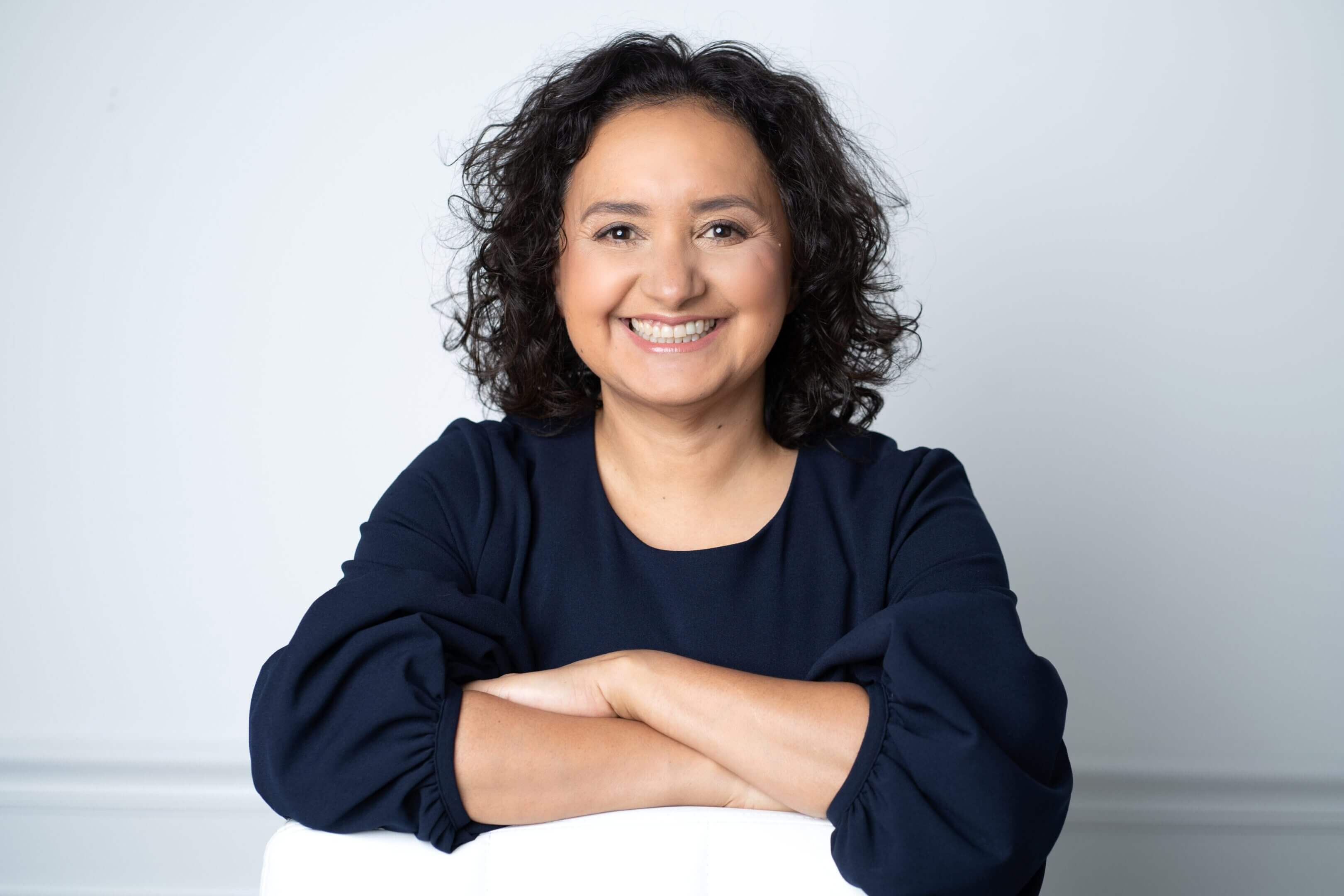 Smiling woman with curly hair in a dark sweater posing with crossed arms.
